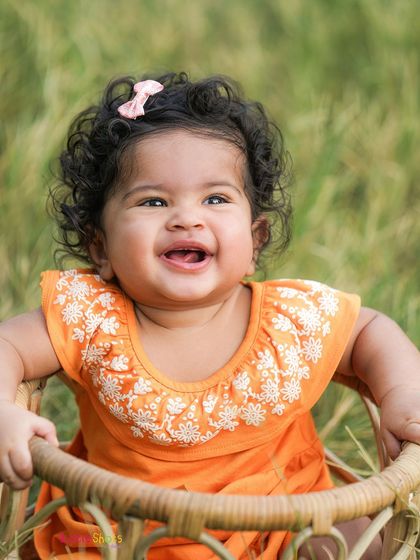 A close-up of pure happiness. This baby girl's radiant smile and bright orange dress make for a warm and joyful outdoor portrait.