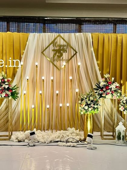 An elegant and classic stage for a 75th birthday celebration. The decor features a golden pleated backdrop, soft white drapes, and beautiful floral arrangements.