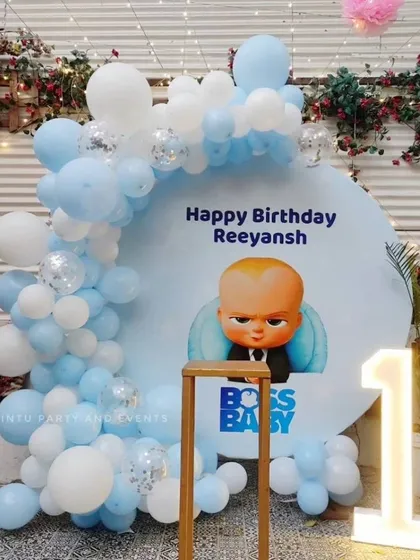A Boss Baby themed first birthday party. This setup includes a custom circular backdrop with the Boss Baby character, a light-up marquee number '1', and a balloon arch in baby blue and white.
