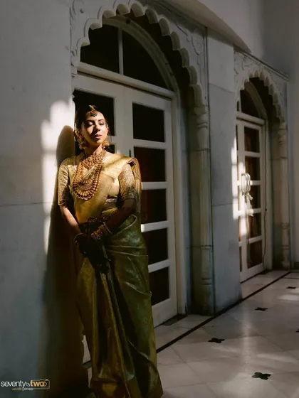 A bride in a golden saree stands in a sunlit corridor, the strong shadows from the arches creating a striking and artistic composition.