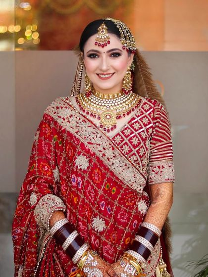 A smiling bride in a traditional red bandhani saree. Her makeup is classic, with a red bindi, gold jewelry, and a warm, happy glow.