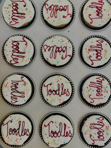 A tray of our vanilla buttercream cupcakes decorated for a corporate farewell party. Each cupcake is topped with the fun message "Toodles" and colorful sprinkles for a sweet and cheerful send-off.