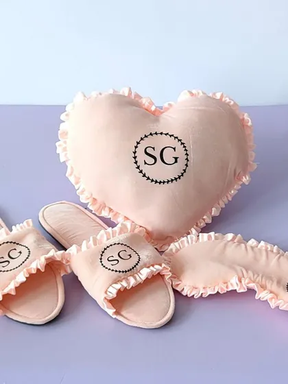 A personalized comfort set for your bridesmaids, featuring a monogrammed heart-shaped pillow, slippers, and eye mask in a soft pink fabric.