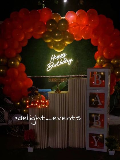 A striking heart-shaped balloon arch in bold red and gold creates a dramatic and romantic backdrop. This setup, complete with a neon sign, 'BABY' blocks, and pedestals, is perfect for celebrating a loved one.