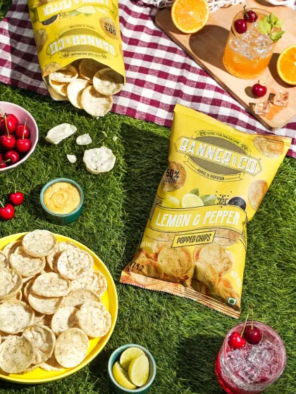 A sunny picnic flatlay featuring Banner & Co Lemon & Pepper chips, fresh cherries, and cool drinks. The bright lighting and checkered mat create a cheerful and appetizing summer scene.