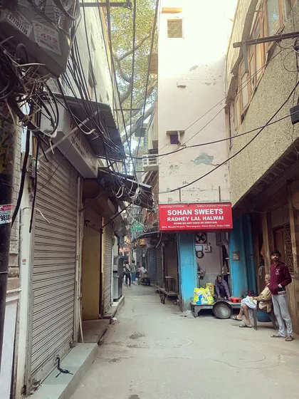 A typical narrow lane in Old Delhi, home to legendary sweet shops like 'Sohan Sweets Radhey Lal Halwai'. We guide you through these historic alleyways to uncover their delicious secrets.