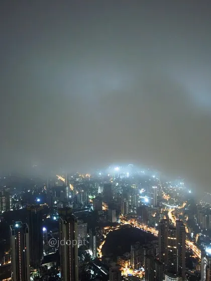 An aerial view of Mumbai's glowing city lights being enveloped by a thick layer of fog and clouds at night.