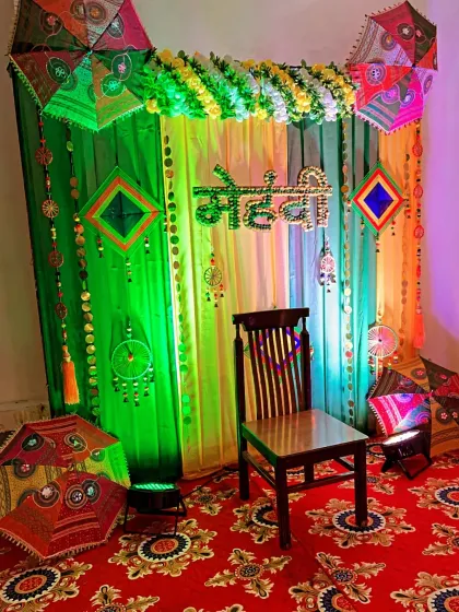 A vibrant and traditional backdrop for a Mehndi ceremony at home. The setup uses colorful drapes in green and yellow, decorated with Rajasthani umbrellas, marigold flower garlands, and ethnic hangings to create a festive atmosphere.