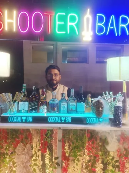 A frontal view of a beautiful outdoor shooter bar setup. The bar is decorated with hanging flowers and features a prominent neon sign and a fully stocked cocktail station.
