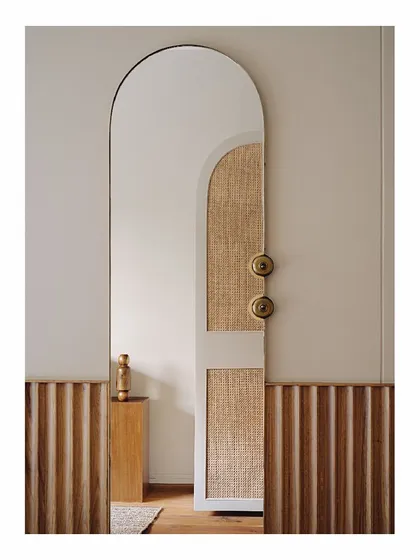 The mother's bedroom features a hand-carved oak wood headboard and wardrobes wrapped in sand-stained wood and cane. The arched mirror and custom totem-like floor lamp, made from plywood offcuts, add a unique, geometric elegance to the space.
