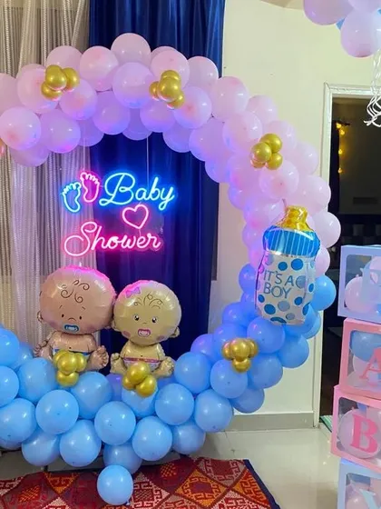 A crescent-shaped balloon arrangement for a "Baby Shower" or gender reveal. This setup uses pink and blue balloons, a neon sign, and baby-themed foil balloons.