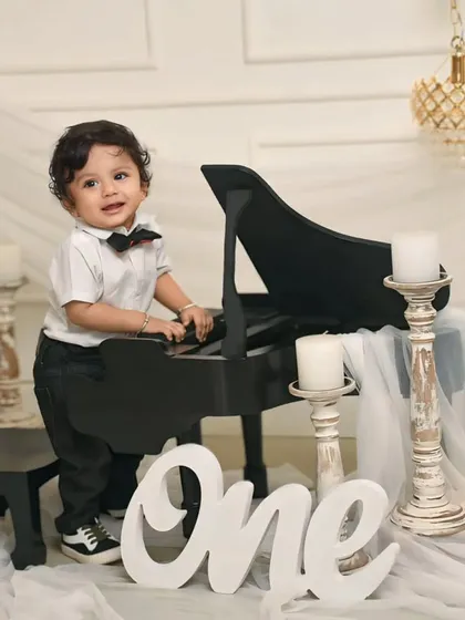This little gentleman is celebrating his first birthday with a classic piano-themed setup. Dressed in a dapper outfit with a bowtie, he creates a charming and sophisticated birthday portrait.