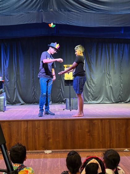 A magician performs a levitation trick for a student volunteer on stage, with the audience watching intently from their seats.