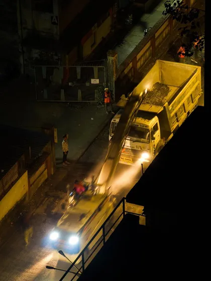 A top-down night shot of a construction site, where a dump truck is being loaded under bright lights, with motion blur from a passing vehicle.
