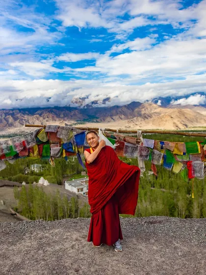 A Buddhist nun in red robes stands on a hilltop in Ladakh, with prayer flags and the vast mountain range behind her. The image conveys a sense of freedom and spiritual devotion.