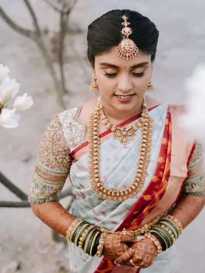 A serene portrait of the bride standing amongst white flowers. Her traditional white saree and gold jewelry create a look of pure elegance and tranquility.