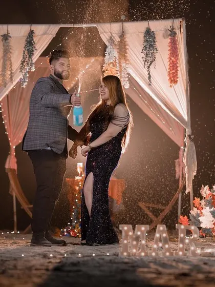A fun and celebratory moment from a "Marry Me" setup on the beach at night, complete with champagne spray and romantic lighting.