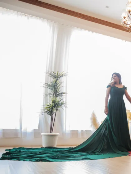 A stunning solo portrait of the bride in her flowing green gown, standing in a brightly lit room. The long train spreads across the floor, creating a dramatic and elegant effect.
