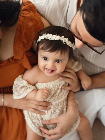 A baby girl in a lace romper sticks her tongue out playfully while being held by her parents. A perfect candid moment from a sitter session.