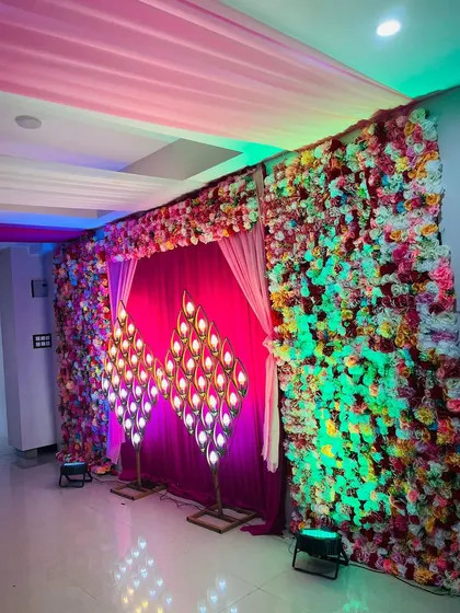 A vibrant stage backdrop for a Sangeet or reception. This setup features a wall of colorful flowers and unique peacock-feather-shaped lamps.