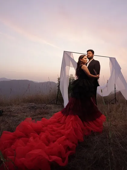 A couple embraces on a hillside at dusk, with a dramatic red and black gown spread out on the ground, creating a powerful and stylish portrait.