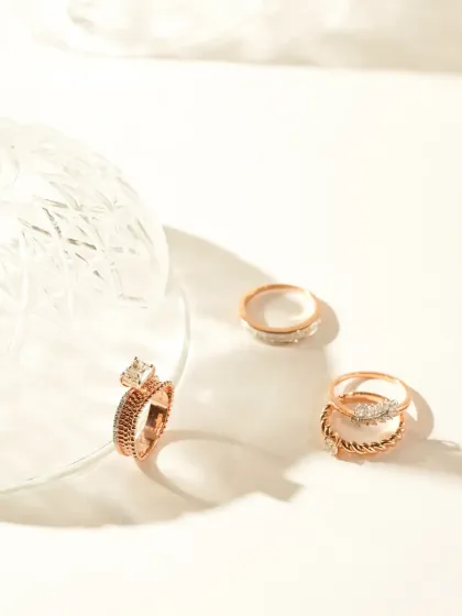 A collection of rose gold rings styled with a crystal glass against a clean, sunlit background. The warm tones and hard shadows create a modern, chic aesthetic that highlights the contemporary design of the rings.