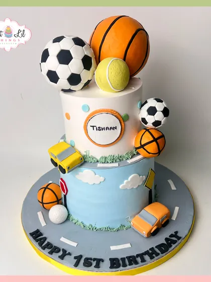 A sporty first birthday cake for a little champ. This two-tiered design is decorated with fondant sports balls like a basketball and soccer ball, as well as little cars.
