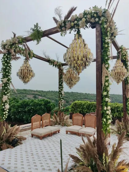 A modern rustic mandap that blends nature with luxury. This outdoor wedding setup features a wooden structure adorned with white roses and greenery, with elegant crystal chandeliers hanging above, all set against a beautiful natural backdrop.