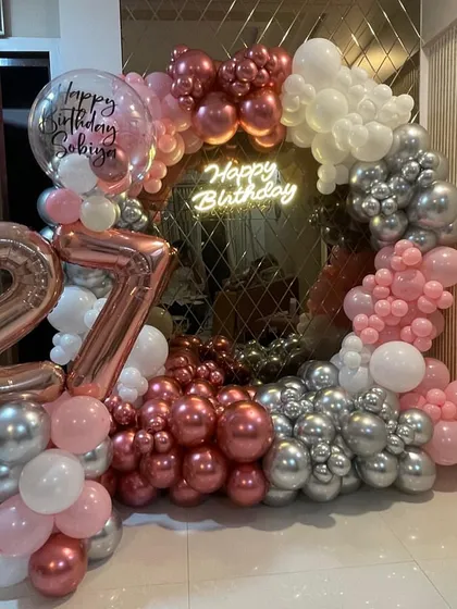 A stylish 27th birthday party with a ring mirror backdrop. The decor is framed by a lush arch of chrome rose gold, pink, and silver balloons, and includes a personalized bubble balloon.