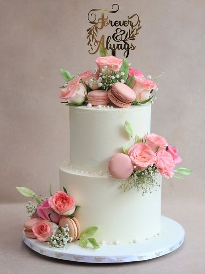 Classic, romantic, and timeless. This two-tiered white buttercream cake is elegantly decorated with fresh pink roses and matching macarons, perfect for an engagement, anniversary, or intimate wedding celebration.