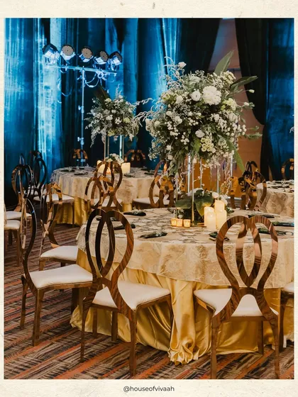An elegant dining table setup with sculptural wooden chairs, a gold and ivory tablecloth, and a tall white floral centerpiece, perfect for a luxurious soiree.