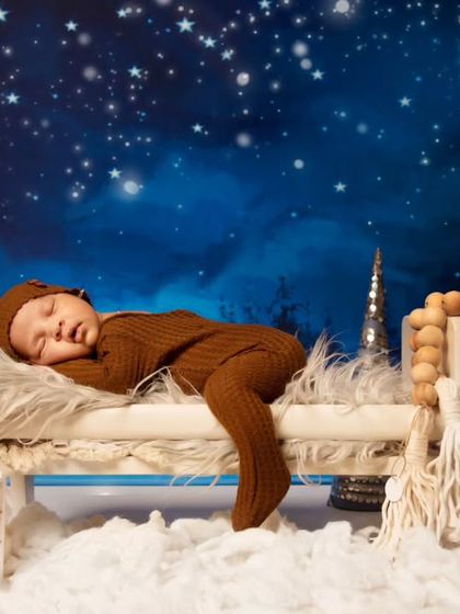 This little one is stretched out and sleeping on a miniature wooden bed, surrounded by a wintery, star-lit forest scene. It’s a perfect example of a cozy and imaginative newborn portrait.