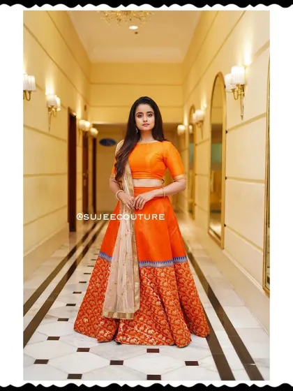 A full-length shot of Chaitra Vasudevan in her custom orange lehenga. The simple, elegant blouse is paired with a voluminous skirt featuring a traditional Benarasi weave, creating a balanced and timeless look.