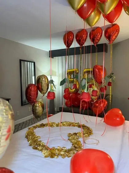 An elegant hotel room decoration for a 55th birthday, using red and gold balloons, heart-shaped foils, and a heart made of petals on the bed.