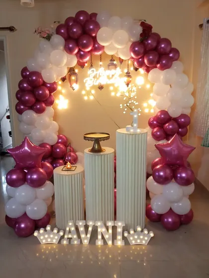 An elegant birthday decoration with a balloon arch in magenta and white. The setup is beautifully lit with hanging lanterns and a light-up name display.
