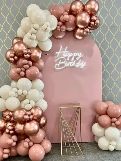 A stylish birthday setup with a pink arch backdrop. The organic balloon garland in rose gold and white, paired with a neon sign, creates a modern and feminine look.