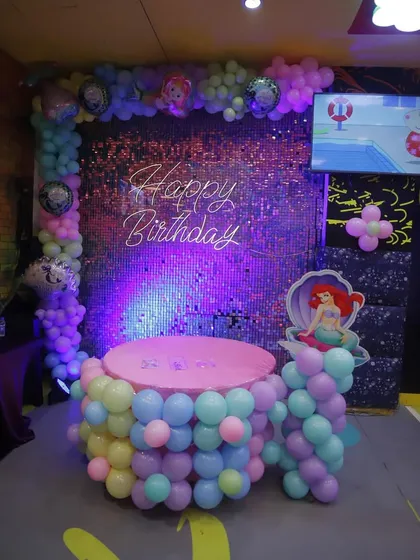 A mermaid-themed birthday party setup featuring characters from The Little Mermaid. The pastel balloon columns and shimmering backdrop create an under-the-sea vibe.