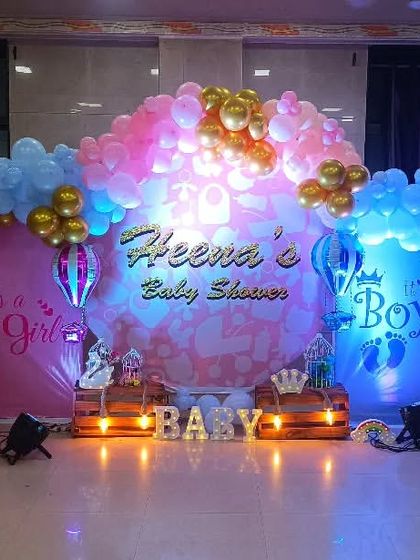 A bright and colorful baby shower setup with a gender theme. The stage is lit with pink and blue lights and features a large balloon arrangement with gold accents.