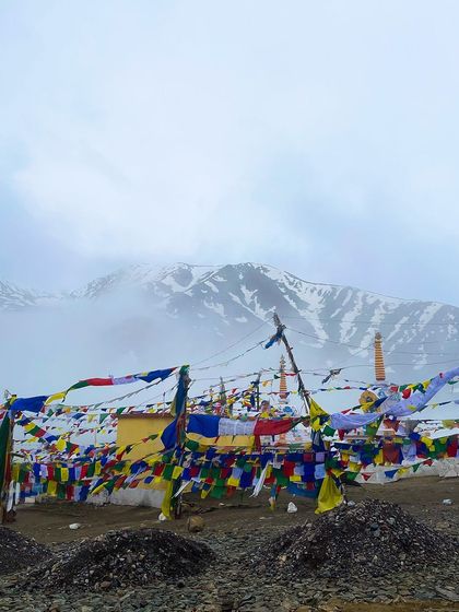 Buddhist prayer flags flutter in the wind at a high mountain pass in Spiti. The misty, snow-covered peaks in the background create a sense of peace and spiritual elevation.