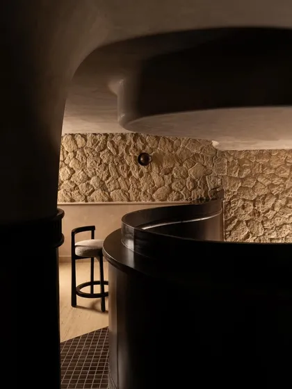 A corridor view, highlighting the contrast between the rough stone wall and the smooth, dark wood of the bar. This passage is a sensory transition, a moment of pause in the spatial narrative.