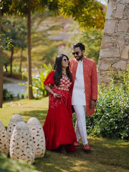 A candid walking shot in a lush garden setting in Rajasthan. The couple's vibrant red traditional outfits stand out beautifully against the greenery.