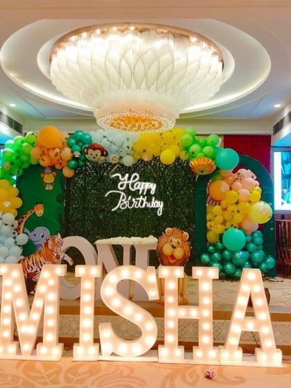 A large jungle-themed stage in a banquet hall, featuring a green foliage backdrop, animal-themed balloon clusters, and oversized light-up letters spelling the birthday girl's name.