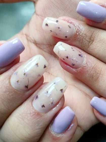 A stylish manicure combining solid pastel purple nails with accent nails featuring dried flowers embedded in a sheer base.