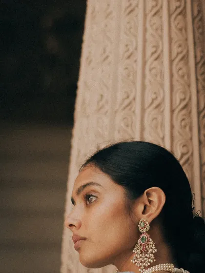 A close-up profile of the bride, her gaze thoughtful and serene. The intricate details of the stone pillar beside her complement the elegance of her jewelry. This is a quiet, artful bridal portrait.