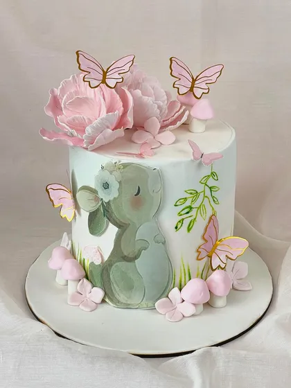 A whimsical hand-painted cake featuring a cute bunny surrounded by pink sugar flowers, mushrooms, and delicate butterflies.