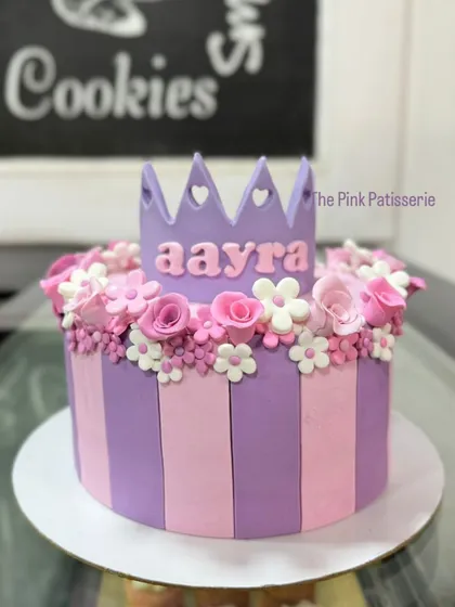 A close-up of our popular princess crown cake. The vertical pink and purple stripes are made from fondant, and the cake is topped with a floral crown.