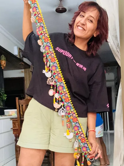 This is me with my 'TAMAARA' toran, a piece that is pure joy to create. It’s a celebration of beauty and mindful artistry, designed to light up your space with color and soul. Each element, from the embroidered lace to the wooden birds and lotuses, is hand-assembled by me.