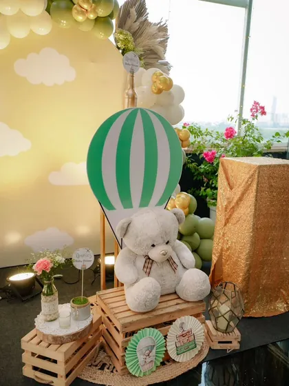 A cozy corner at the travel-themed baby shower. A large teddy bear sits on wooden crates next to a hot air balloon prop, creating a sweet photo opportunity for guests.