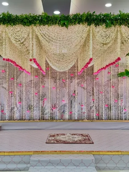 This stage was designed for a modern Janoi ceremony with a serene pink and white color palette. The backdrop is a curtain of delicate white floral strings accented with pink flowers, framed by traditional banana trees, blending tradition with contemporary aesthetics.