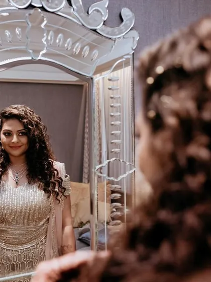 A bride looking at her reflection. Her curly hair is styled beautifully, and she's wearing an elegant embellished gown, ready for her reception.